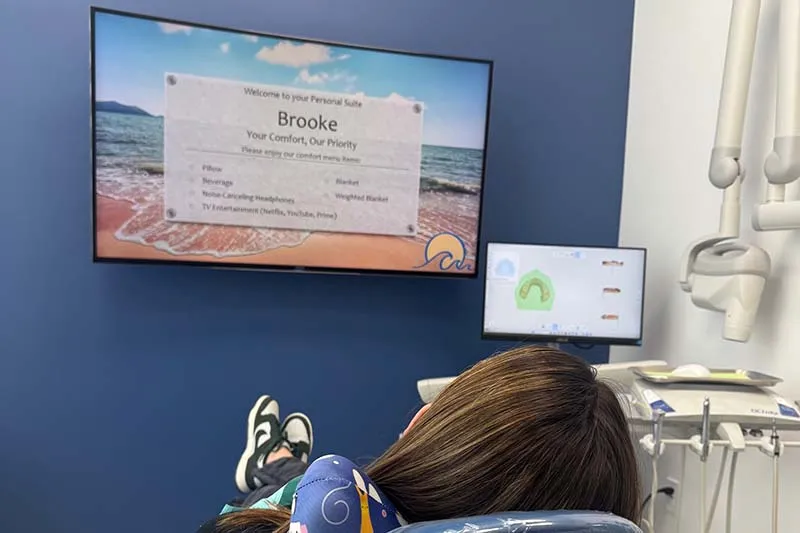 Paciente mirando la televisión mientras está en el sillón de examen.. El televisor muestra todas las comodidades disponibles para ella, incluidas: una almohada, bebidas, manta ponderada, manta normal, y la elección del canal de televisión.